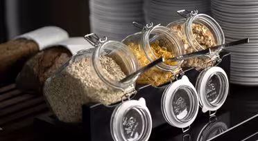 Tre glasburkar fyllda med havre, cornflakes och blandade korn är snyggt arrangerade med skedar i en hållare. De ställs framför en bunt vita tallrikar, bredvid ett bröd på en skärbräda.