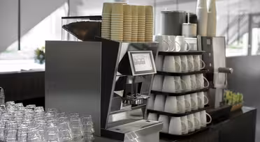 Modern kaffemaskin omgiven av staplade pappersmuggar, porslinsmuggar och plastglas i en kafémiljö. Bänkskivan rymmer även sockerpaket och servettdispensrar.