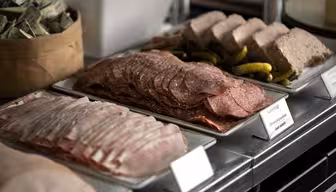 Ett urval av skivade delikatesser, inklusive salami och en annan sort, är snyggt arrangerad på brickor. Pickles placeras bredvid, och det finns små skyltar framför varje bricka. En korg med kryddor eller paket syns i bakgrunden.