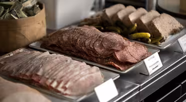 Ett urval av skivade delikatesser, inklusive salami och en annan sort, är snyggt arrangerad på brickor. Pickles placeras bredvid, och det finns små skyltar framför varje bricka. En korg med kryddor eller paket syns i bakgrunden.