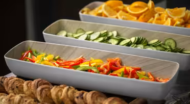 En buffédisplay med tre rektangulära brickor fyllda med skivade apelsiner, gurkor och en blandning av röd, gul och grön paprika. I förgrunden är bakverk upplagda på ett fat.