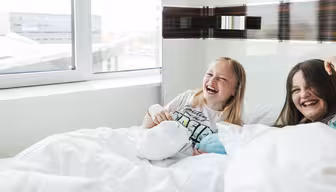 Två flickor skrattar och sitter tillsammans på en säng, täckt med vita filtar, nära ett stort fönster. Rummet är ljust med naturligt ljus som strömmar in. De är båda vända mot kameran och njuter av en lekfull stund.