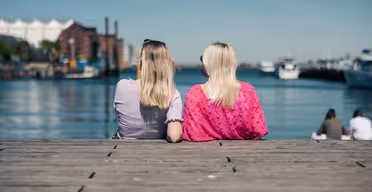 Två individer sitter på en träbrygga vänd mot en lugn vattenmassa med båtar i bakgrunden. Båda har långt hår; en bär en lavendel topp, och den andra bär en rosa blus. Himlen är klar och blå.