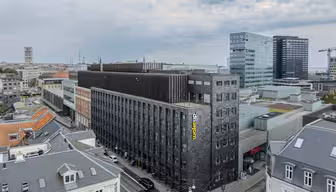 Flygfoto över en stadsbild med en modern, mörkfärgad hotellbyggnad med namnet "Wakeup" på sidan. De omgivande byggnaderna inkluderar äldre byggnader med gråa tak, moderna glasbyggnader och delvis molnig himmel.