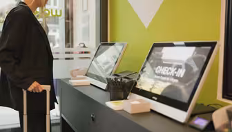 En person står vid en incheckningsdisk på hotellet och använder självbetjäningskiosker. Kioskerna visar ordet CHECK-IN på sina skärmar. Rummet har modern design med limegröna och svarta detaljer. Personen håller ett handtag i en resväska.