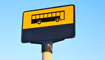 En busshållplatsskylt med gul bakgrund och en svart bussikon överst. Den nedre delen av skylten är svart. Skylten är monterad på en stolpe mot en klarblå himmel.
