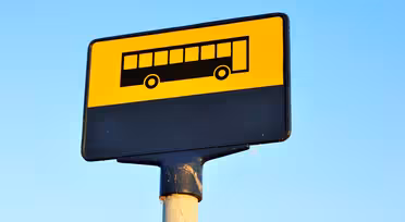 En busshållplatsskylt med gul bakgrund och en svart bussikon överst. Den nedre delen av skylten är svart. Skylten är monterad på en stolpe mot en klarblå himmel.