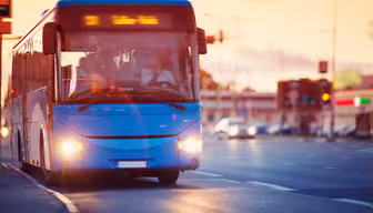 En blå stadsbuss åker nerför en gata i solnedgången, med en suddig bakgrund av byggnader och bilar. Bussen har upplysta destinationsskyltar och strålkastare på, med några passagerare synliga inuti.