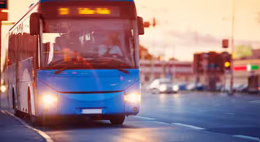 En blå stadsbuss åker nerför en gata i solnedgången, med en suddig bakgrund av byggnader och bilar. Bussen har upplysta destinationsskyltar och strålkastare på, med några passagerare synliga inuti.