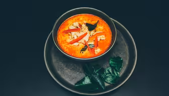 En skål med röd curry med skivad röd paprika och garnering sitter på en mörk tallrik. Färska gröna blad är arrangerade på tallriken bredvid skålen. Curryn verkar krämig och rikt färgad.