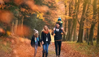 En familj på fyra går genom en skog på hösten. Föräldrarna ler, håller hand, med pappan som bär ett barn på sina axlar. Det andra barnet går bredvid, omgivet av livfulla röda och orange löv.