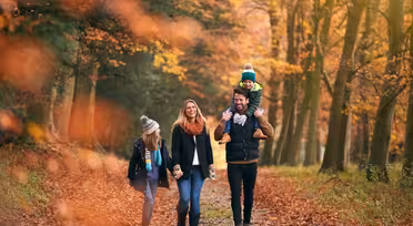 En familj på fyra går genom en skog på hösten. Föräldrarna ler, håller hand, med pappan som bär ett barn på sina axlar. Det andra barnet går bredvid, omgivet av livfulla röda och orange löv.