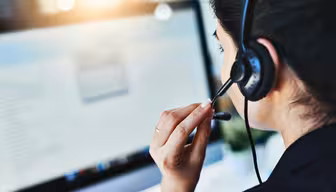 En person som bär ett headset sitter framför en datorskärm. Skärmen visar suddig text och grafik. Scenen föreslår ett callcenter eller kundsupportinställning med fokus på kommunikation.