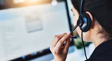 En person som bär ett headset sitter framför en datorskärm. Skärmen visar suddig text och grafik. Scenen föreslår ett callcenter eller kundsupportinställning med fokus på kommunikation.