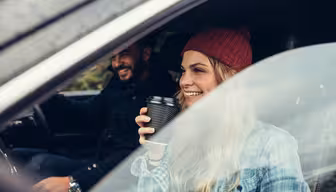 En kvinna i en röd mössa och rutig skjorta sitter i en bil och ler och håller i en kaffekopp. En man sitter bredvid henne och ler också. De ser ut att njuta av en biltur en trevlig dag, med utsikt genom bilfönstret.
