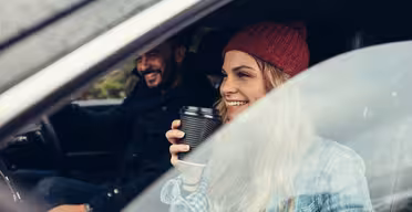 En kvinna i en röd mössa och rutig skjorta sitter i en bil och ler och håller i en kaffekopp. En man sitter bredvid henne och ler också. De ser ut att njuta av en biltur en trevlig dag, med utsikt genom bilfönstret.