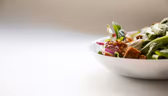 En närbild av en skål med sallad som innehåller bladgrönsaker, rödlök, linser och krutonger. Skålen är placerad på höger sida mot en minimalistisk, neutral bakgrund.