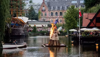 En träskulptur i brand flyter på en kanal med människor som tittar på från båtar och närliggande kaféer. En byggnad i rött tegel och träd omger området, och ett högt skepp är dockat till vänster. En flagga är synlig till höger.