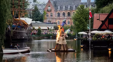 En träskulptur i brand flyter på en kanal med människor som tittar på från båtar och närliggande kaféer. En byggnad i rött tegel och träd omger området, och ett högt skepp är dockat till vänster. En flagga är synlig till höger.
