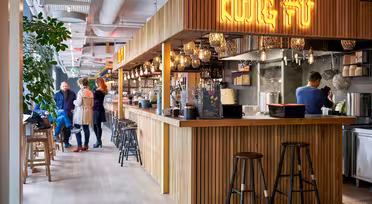 En modern restauranginredning med träbänk och pallar. På en neonskylt står det "KUNG FU". Hänglampor lyser upp utrymmet. Gästerna sitter och pratar medan personalen lagar mat bakom disken. En lummig växt lägger till grönska till scenen.