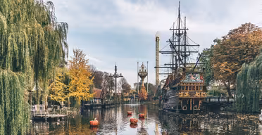 En vacker utsikt över en nöjesparksjö med ett dockat piratskepp. Flytande pumpor är i vattnet, omgivna av höstfärgade träd. I bakgrunden reser sig torn och attraktioner mot en molnig himmel.