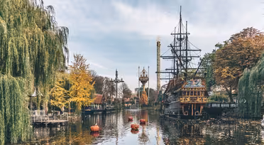 En vacker utsikt över en nöjesparksjö med ett dockat piratskepp. Flytande pumpor är i vattnet, omgivna av höstfärgade träd. I bakgrunden reser sig torn och attraktioner mot en molnig himmel.