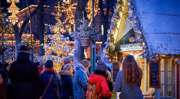 En festlig utomhusjulmarknadsscen med människor samlade runt ett dekorerat stånd. Varma lampor och hängande lyktor lyser upp området. Ett snötäckt tak förhöjer den mysiga vinterstämningen och träden pryds av glödande ljusslingor.