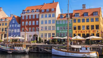 Färgglada byggnader kantar vattnet i Nyhavn, Köpenhamn, med traditionella träbåtar anlagda längs kanalen. Klarröda, gula, gröna och krämfärgade fasader är framträdande under en klarblå himmel.