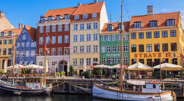 Färgglada byggnader kantar vattnet i Nyhavn, Köpenhamn, med traditionella träbåtar anlagda längs kanalen. Klarröda, gula, gröna och krämfärgade fasader är framträdande under en klarblå himmel.