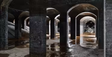 En svagt upplyst, välvd underjordisk vattenreservoar med flera tegelpelare. Golvet är blankt och fuktigt, vilket reflekterar strukturens bågar och pelare. Skuggor skapar en mystisk atmosfär.