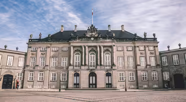 En storslagen, historisk byggnad med en utsmyckad fasad med pelare, statyer och stora fönster. Den har ett mörkt tak och står mot en delvis molnig himmel, med en kullerstensgård framför.