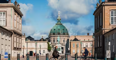 Två personer cyklar på en kullerstensgata mot en stor byggnad med en grön kupol under en blå himmel med moln. Arkitekturen är klassisk, med höga strukturer som flankerar båda sidor av gatan.