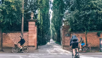 Två cyklister cyklar längs en asfalterad väg bredvid en hög tegelvägg med en ingång som leder till en trädkantad gångväg. Scenen är ljus och omgiven av frodig grönska. En tredje person på cykel syns längre ner på vägen nära infarten.
