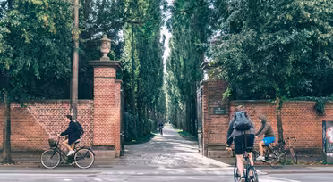 Två cyklister cyklar längs en asfalterad väg bredvid en hög tegelvägg med en ingång som leder till en trädkantad gångväg. Scenen är ljus och omgiven av frodig grönska. En tredje person på cykel syns längre ner på vägen nära infarten.