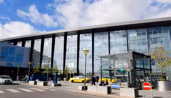Exteriör vy av en flygplatsterminal med en modern glasfasad. Gula taxibilar och en grupp människor står framför. Några träd planteras nära ingången. Blå himmel och moln syns ovanför. Ett övergångsställe och skyltar finns i förgrunden.