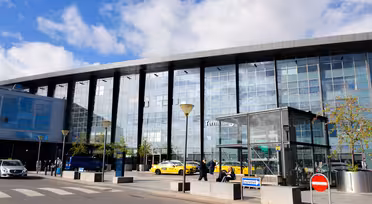 Exteriör vy av en flygplatsterminal med en modern glasfasad. Gula taxibilar och en grupp människor står framför. Några träd planteras nära ingången. Blå himmel och moln syns ovanför. Ett övergångsställe och skyltar finns i förgrunden.