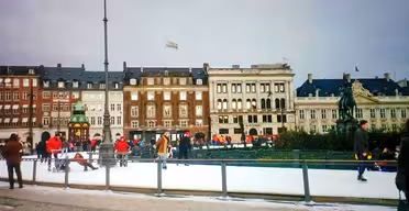 Människor åker skridskor på en rink på ett stadstorg med historiska byggnader i bakgrunden. Scenen innehåller en staty och myllrar av skridskoåkare och åskådare omgiven av en vintrig atmosfär.
