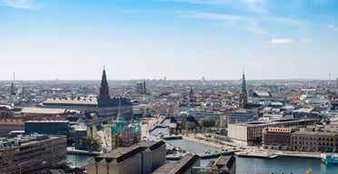 Flygfoto över en europeisk stad med historiska och moderna byggnader, en central flod och flera spiror. Himlen är klar med några moln, vilket förstärker det panoramiska stadslandskapet.