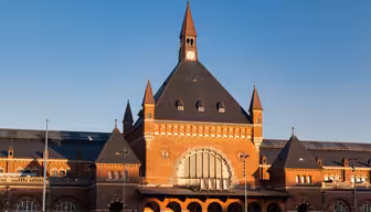 Historisk tegeljärnvägsstation med ett stort välvt fönster, omgivet av mindre spiror, under en blå himmel. Byggnaden har en central klocka och ett spetsigt tak, som frammanar arkitektur från början av 1900-talet.