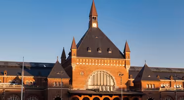 Historisk tegeljärnvägsstation med ett stort välvt fönster, omgivet av mindre spiror, under en blå himmel. Byggnaden har en central klocka och ett spetsigt tak, som frammanar arkitektur från början av 1900-talet.