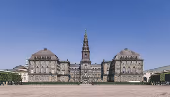 En majestätisk, historisk byggnad med ett centralt torn toppat av en spira, mot en klarblå himmel. Strukturen har symmetriska vingar, stora fönster och stendetaljer, omgiven av en rymlig innergård med klippta häckar.