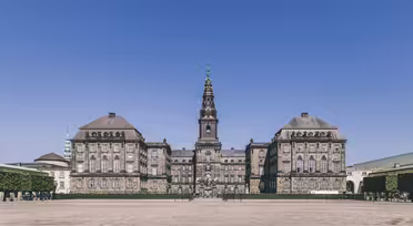 En majestätisk, historisk byggnad med ett centralt torn toppat av en spira, mot en klarblå himmel. Strukturen har symmetriska vingar, stora fönster och stendetaljer, omgiven av en rymlig innergård med klippta häckar.