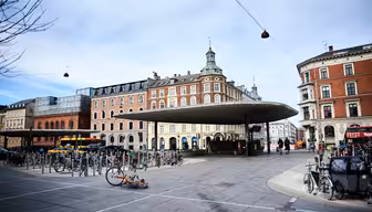 Ett livligt torg med en modern, böjd metallkapell i mitten. Runt torget finns historiska flervåningsbyggnader. Många cyklar står parkerade och folk går i närheten. En cykel ligger på marken. Himlen är delvis molnig.