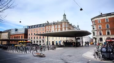 Ett livligt torg med en modern, böjd metallkapell i mitten. Runt torget finns historiska flervåningsbyggnader. Många cyklar står parkerade och folk går i närheten. En cykel ligger på marken. Himlen är delvis molnig.