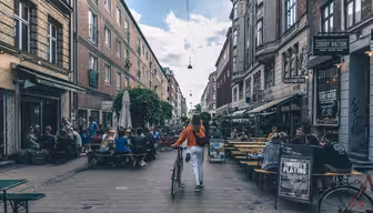 En person som går med en cykel på en livlig gata kantad av tegelbyggnader och uteserveringar. Folk sitter och umgås vid borden. Himlen är delvis molnig och skyltar och grönska pryder området.