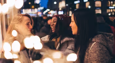 Tre kvinnor ler och pratar i en festlig, upplyst gatumiljö. Scenen är livlig, med suddiga ljus som skapar en varm glöd. Stämningen är glad och livlig med kvällsaktivitet.