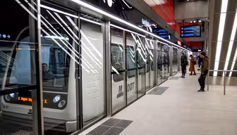 Människor väntar på ankomsten av ett modernt tunnelbanetåg vid en tunnelbanestation med glasbarriärer. Scenen är väl upplyst med digitala skyltar som visar information ovan. Passagerare syns inuti tåget genom stora fönster.