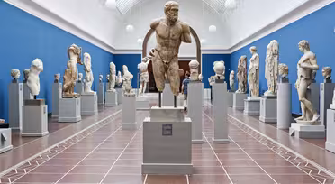 Ett museigalleri med blå väggar visar många klassiska skulpturer på piedestaler. Skulpturerna varierar i storlek och föreställer människofigurer, byster och partiella statyer. En stor framstående staty står centralt. Galleriet har ett välvt glastak.