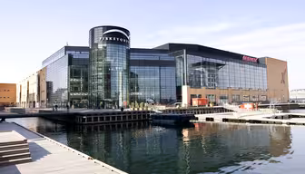 Modernt köpcentrum vid vattnet med stora glasfönster och en cylindrisk entré märkt "Fisketorvet". Omgiven av stilla vatten, trägångar och klar himmel syns byggnadens reflektion i vattnet.