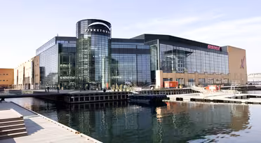 Modernt köpcentrum vid vattnet med stora glasfönster och en cylindrisk entré märkt "Fisketorvet". Omgiven av stilla vatten, trägångar och klar himmel syns byggnadens reflektion i vattnet.
